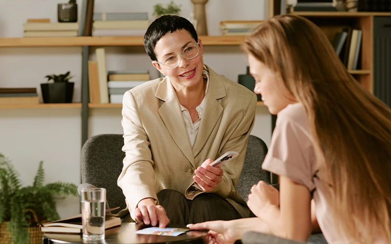 A therapist holding cards talks to a client in a comfortable room with bookshelves.