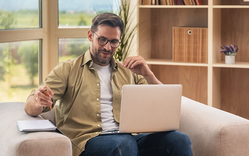 A man sitting on a couch using a laptop with a notebook nearby.
